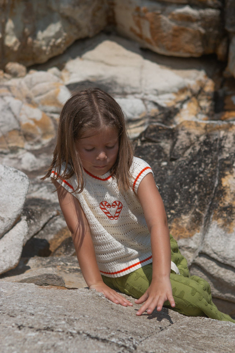 Child wearing a white knit vest with red candy canes and green towelling pants standing on a stone surface.