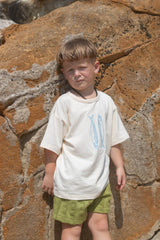 Child wearing a white t-shirt with blue fish and green towelling shorts standing against a rocky wall.