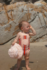 Baby in a striped singletsuit standing on a beach with rocks in the background