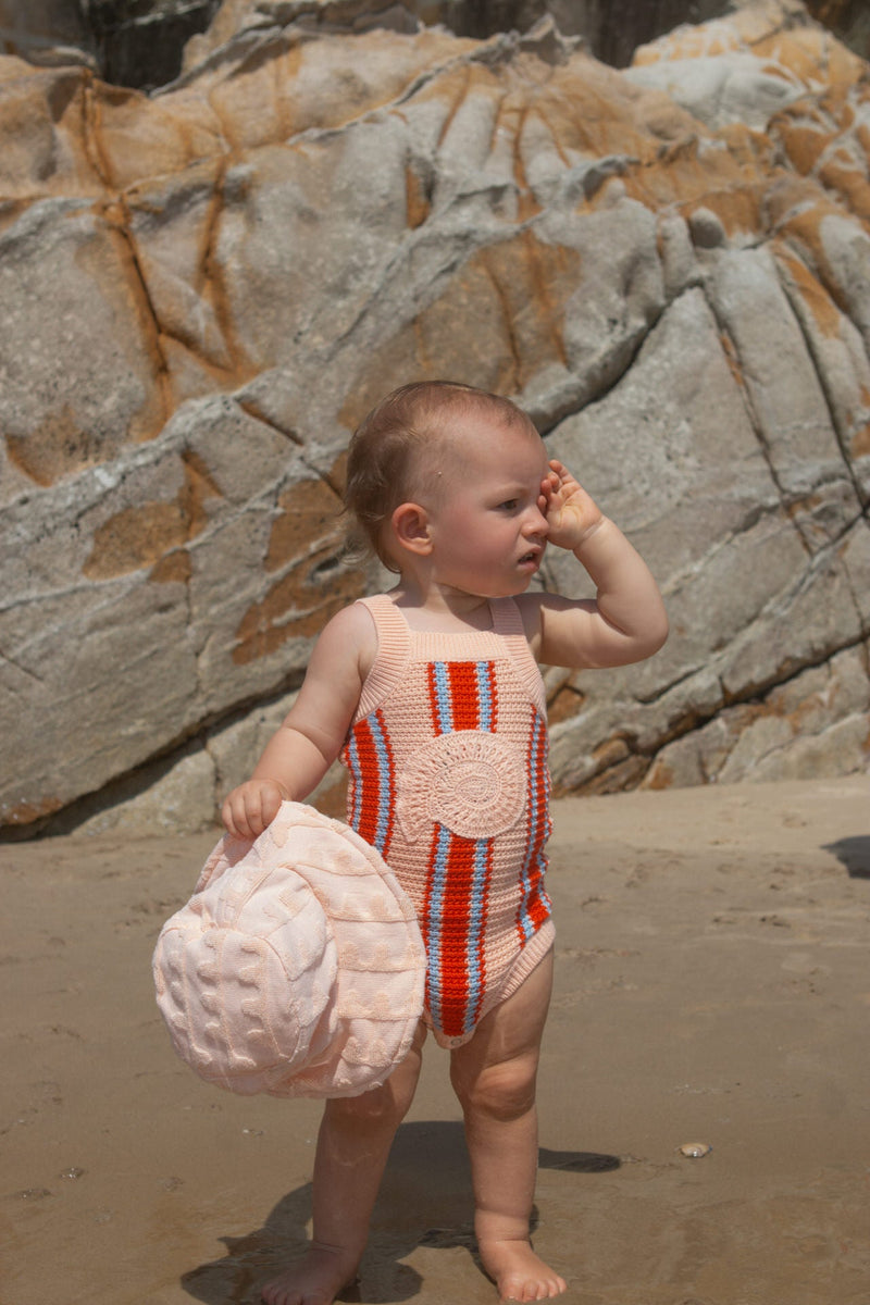 Baby in a striped singletsuit standing on a beach with rocks in the background