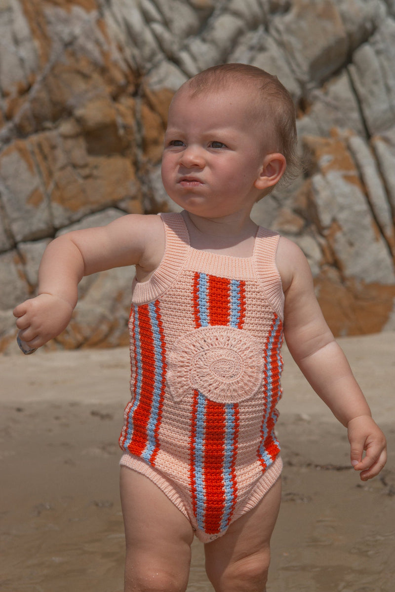 Baby in a striped singletsuit standing on a beach with rocks in the background