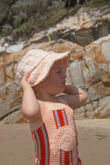 Child wearing a pink sun hat and striped singletsuit on a rocky beach.