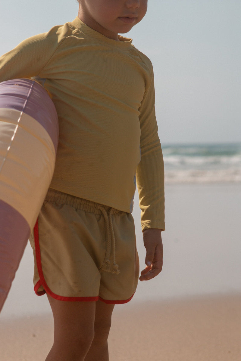 Child in green long-sleeve rash guard and green shorts standing on a beach.